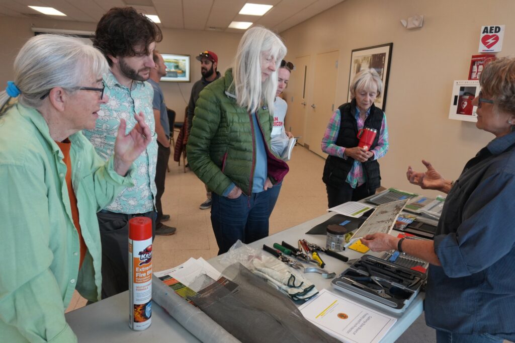 A presenter shows residents ember-resistant vent mesh and home hardening tools laid out on a table